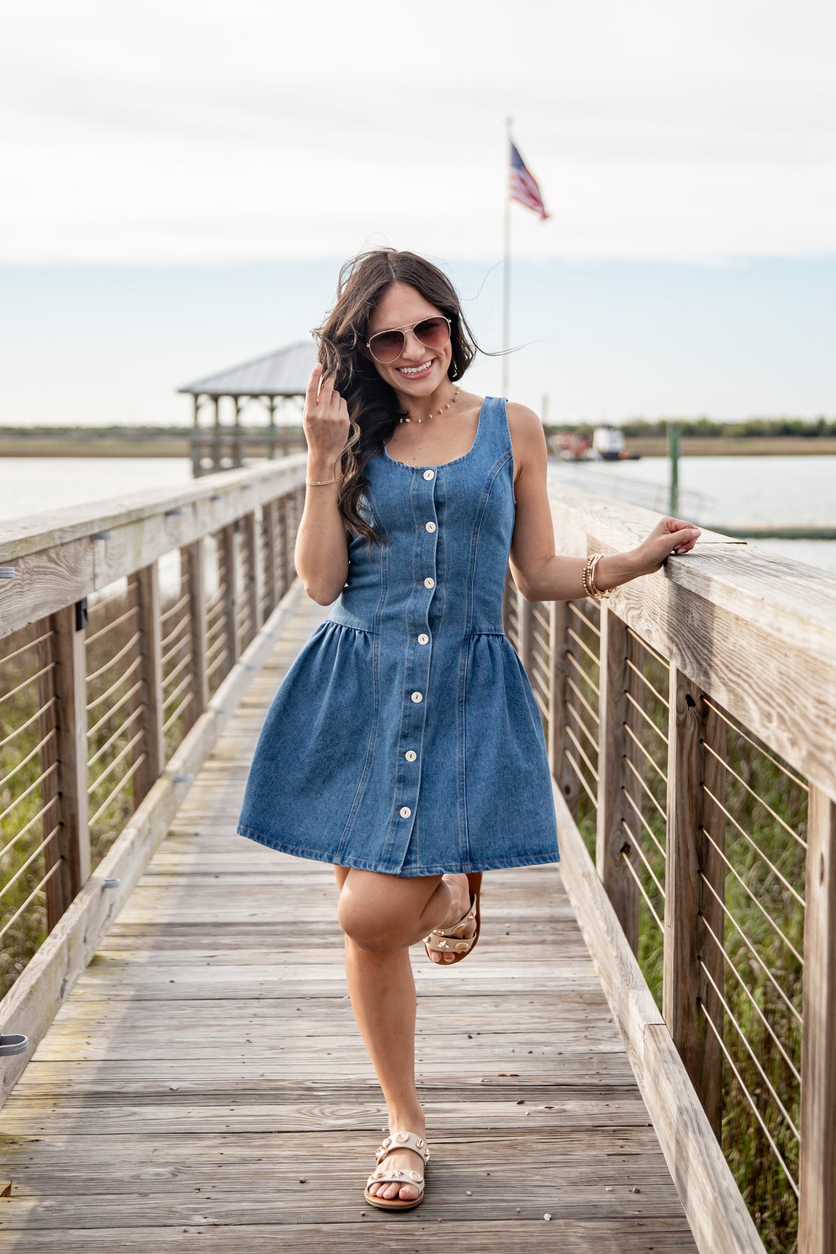 Woman in a blue dress standing on a wooden boardwalk by a waterfront.