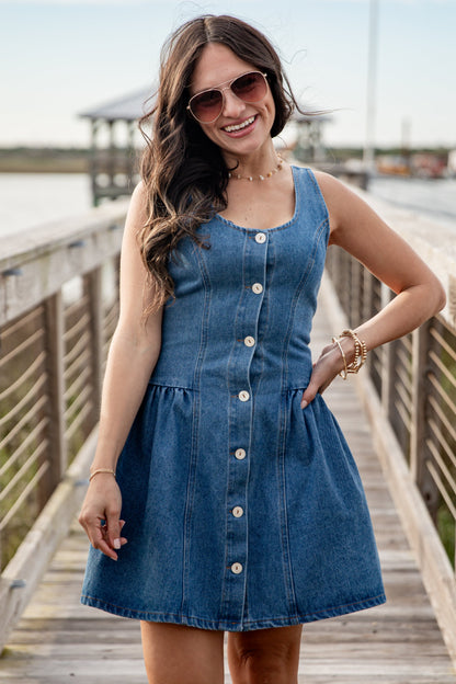Woman wearing a blue denim dress on a wooden dock by a body of water.