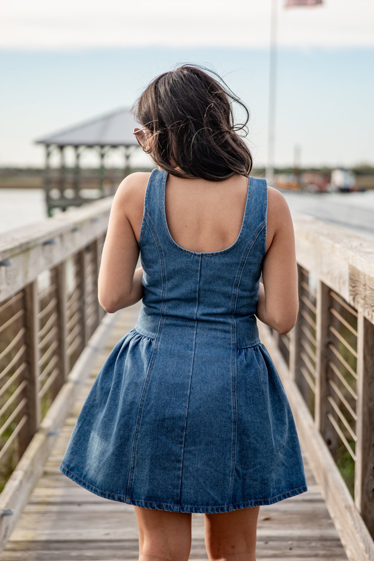 Woman in a blue denim dress standing on a wooden boardwalk by a waterfront.