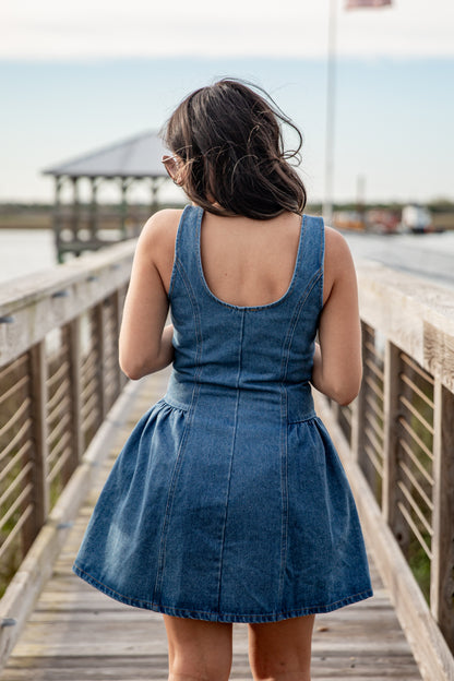 Woman in a blue denim dress standing on a wooden boardwalk by a waterfront.