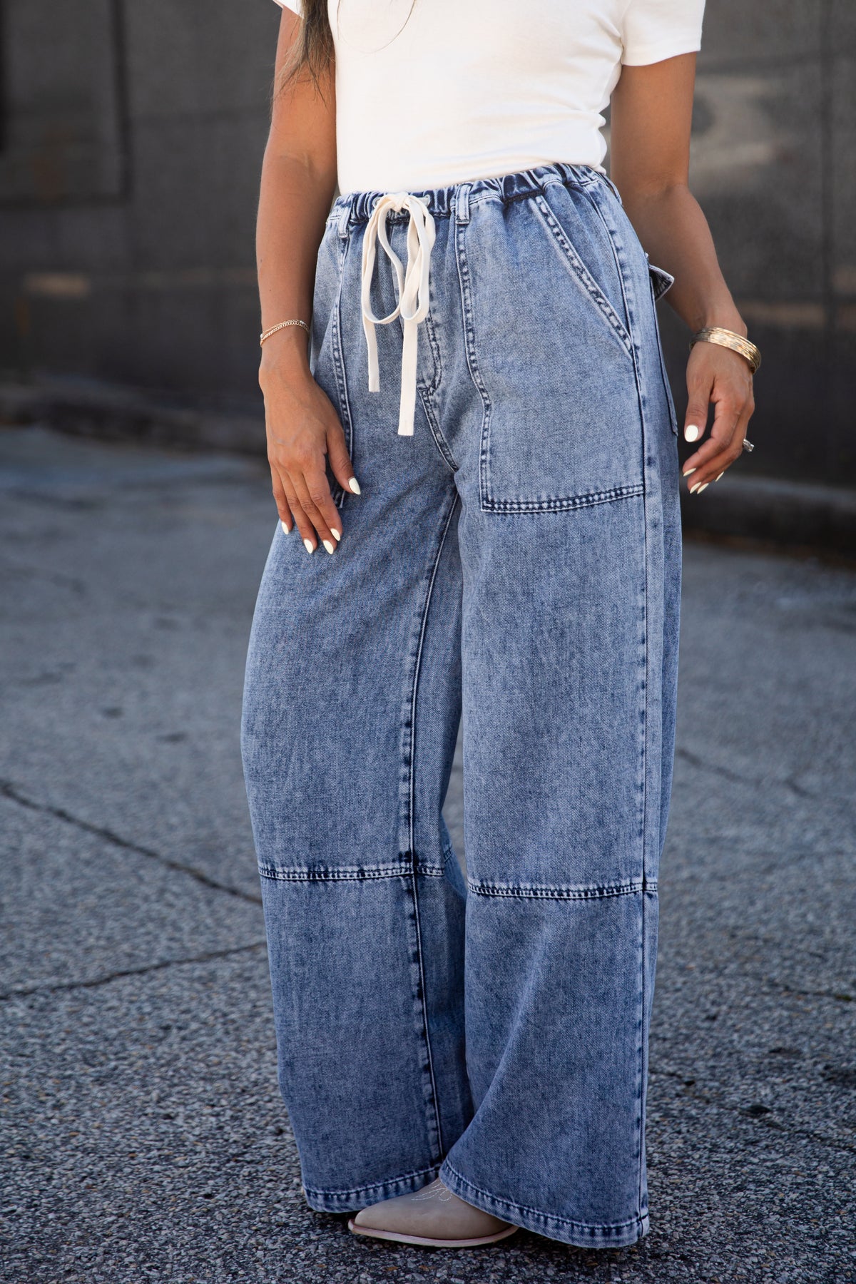 A person wearing wide-leg light blue denim jeans with a white drawstring waist, paired with a white top and gray shoes, standing outdoors on a paved surface.