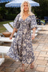 Woman in a floral dress standing on a patio with outdoor furniture and umbrellas in the background.