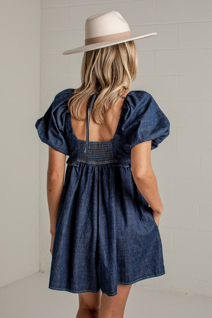 A woman viewed from behind wearing a dark blue denim-like puff-sleeve dress with a smocked back and a cream wide-brim hat, standing against a light neutral wall.