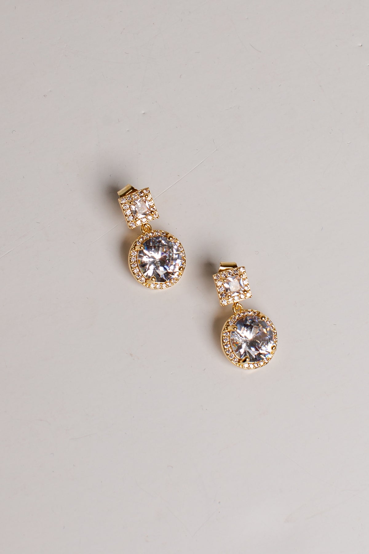 A pair of gold drop earrings with large clear round gemstones and smaller clear stones above them, photographed on a plain light gray background.