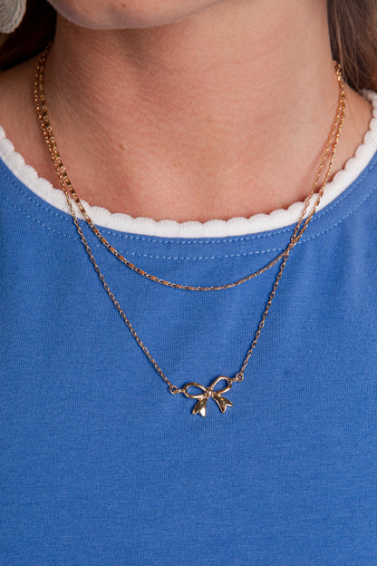 Gold necklace with a bow pendant worn by a person on a blue shirt.