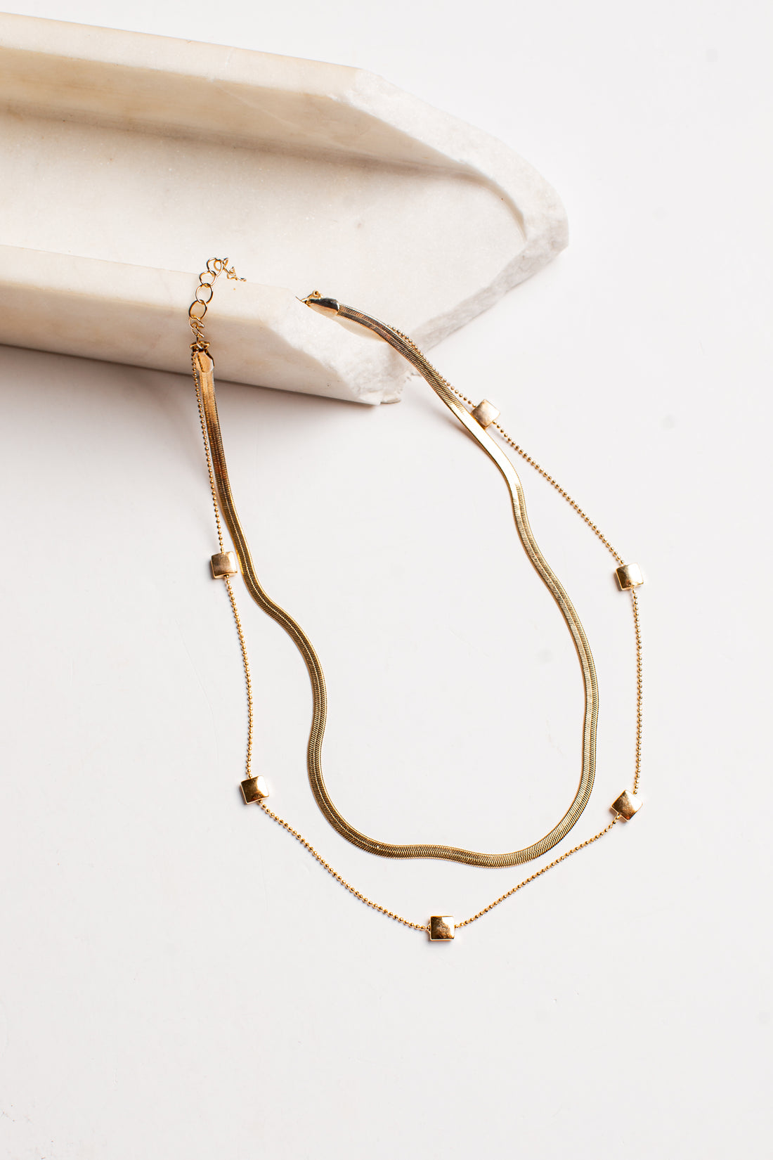 A double-layered gold necklace featuring a textured snake chain and a delicate chain with small gold heart charms, displayed on a light cream-colored wooden block against a white background.