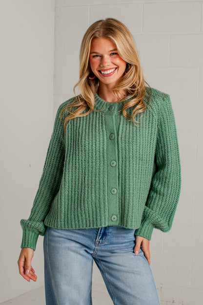 A smiling blonde woman wearing a sage green ribbed knit cardigan with button-front closure and light blue jeans, standing against a light gray brick wall.