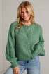 A smiling blonde woman wearing a chunky knit sage green cardigan sweater with button-front closure and light blue jeans, posed against a light gray brick wall.