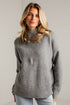A woman wearing a heather gray ribbed knit mock neck sweater and light blue jeans, standing against a light gray brick wall.