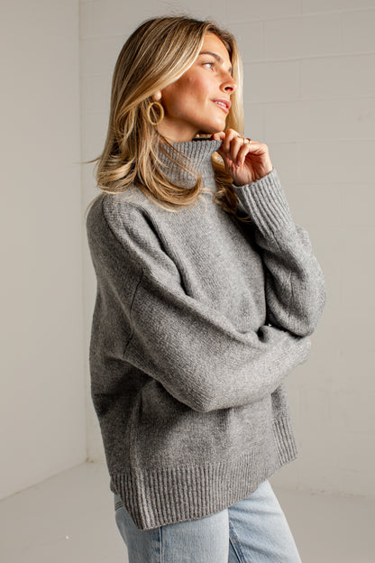 A woman wearing an oversized heather gray ribbed turtleneck sweater and light blue jeans, standing against a light gray wall.