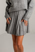 A person wearing a gray ribbed knit sweater and matching pleated mini skirt, standing against a plain light gray wall.