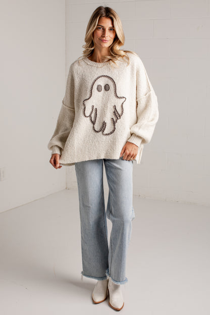A woman modeling a cream-colored knit sweater with a large brown ghost motif on the front, paired with light-wash jeans and white ankle boots, standing against a white brick wall.