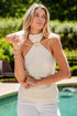 Woman in a white halter top and pants standing by a pool with greenery in the background