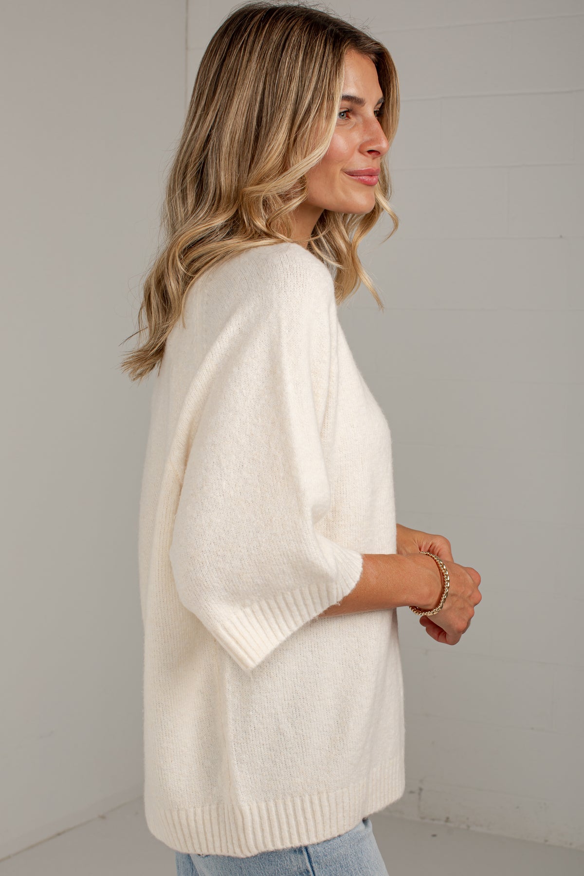 A woman wearing a cream-colored knit sweater with elbow-length sleeves, shown from a side-back angle against a light gray wall.