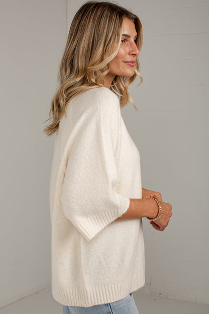 A woman wearing a cream-colored knit sweater with elbow-length sleeves, shown from a side-back angle against a light gray wall.