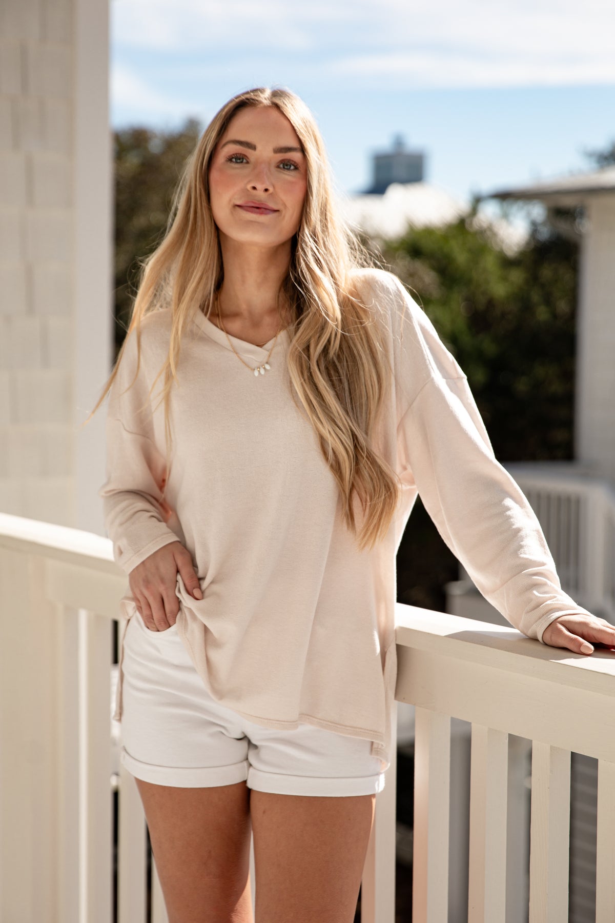 Woman in a beige cardigan and white shorts standing on a balcony with trees and buildings in the background.