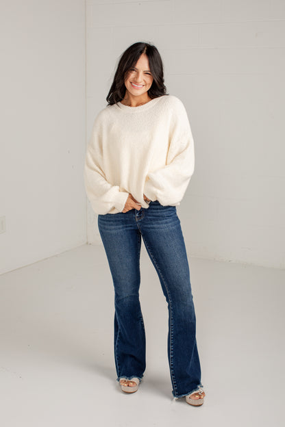 A woman modeling a cream-colored, oversized knit sweater and dark blue flared jeans against a plain white studio background.