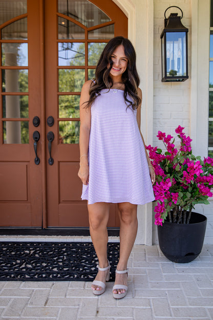 Woman in a purple checkered dress standing on a porch with a brown door and pink flowers.