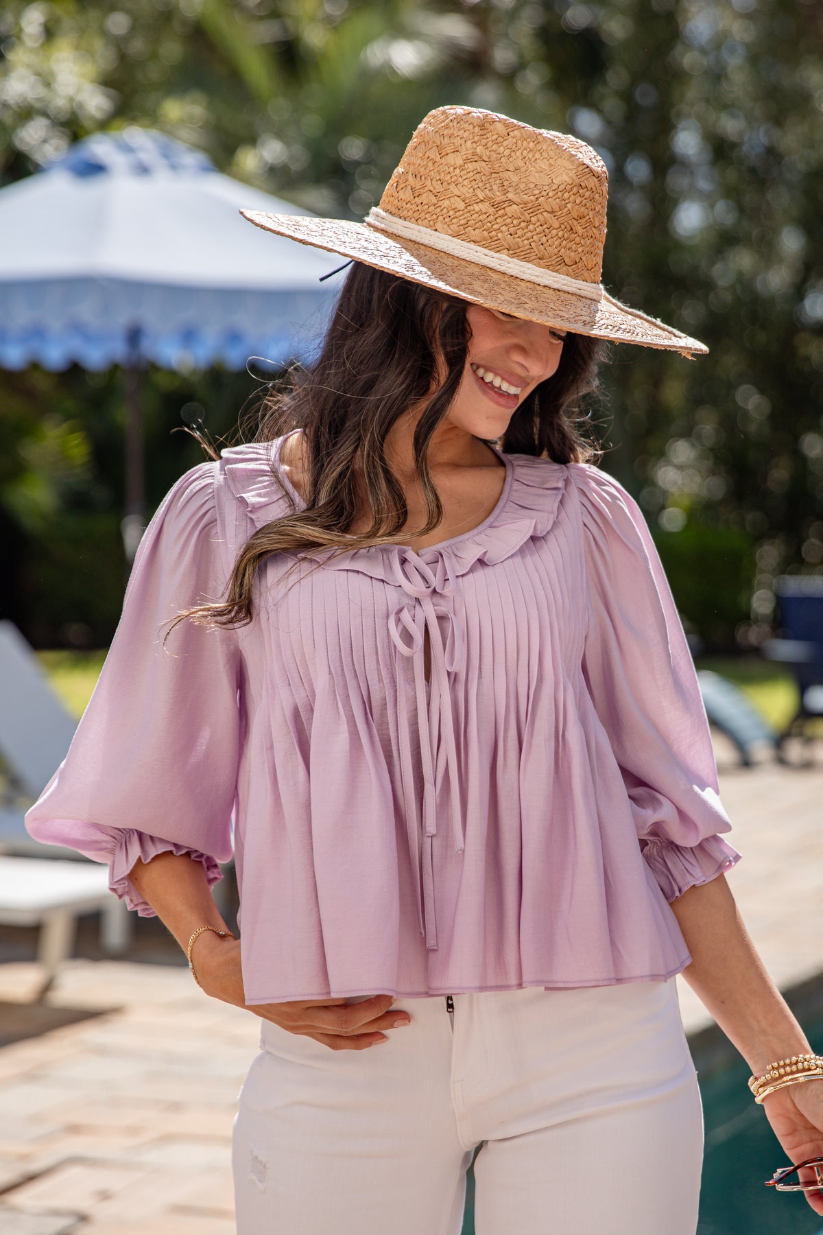 Woman wearing a straw hat and light purple blouse with white pants outdoors.