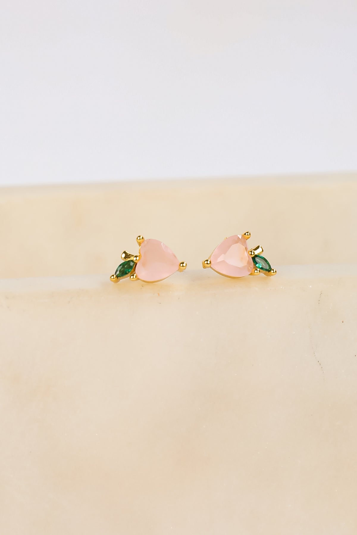 Pair of pink heart-shaped earrings with green gemstones on a beige surface.