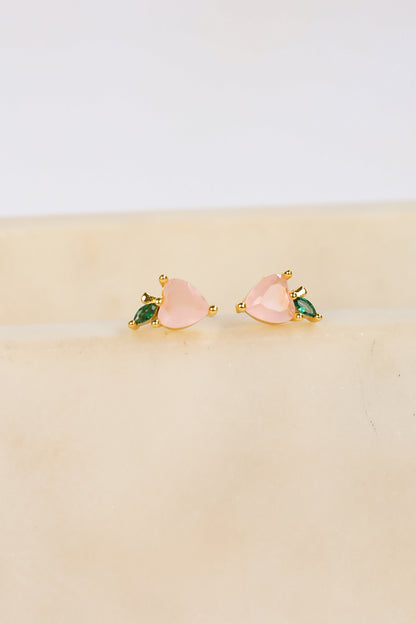 Pair of pink heart-shaped earrings with green gemstones on a beige surface.
