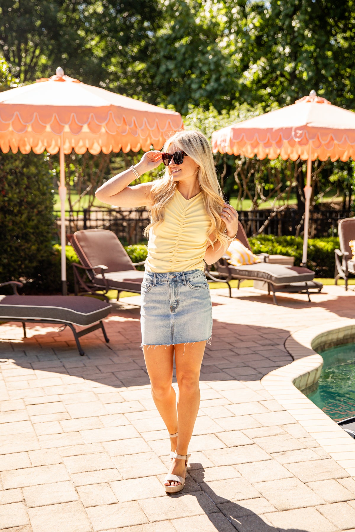Woman in a yellow top and denim skirt standing by a pool with orange umbrellas and lounge chairs.