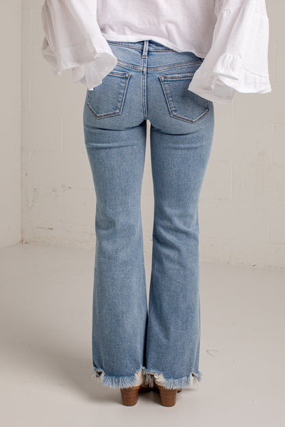 Rear view of a person wearing light-wash blue flare jeans with a frayed hem, paired with a white blouse and brown heels, standing against a plain off-white wall.