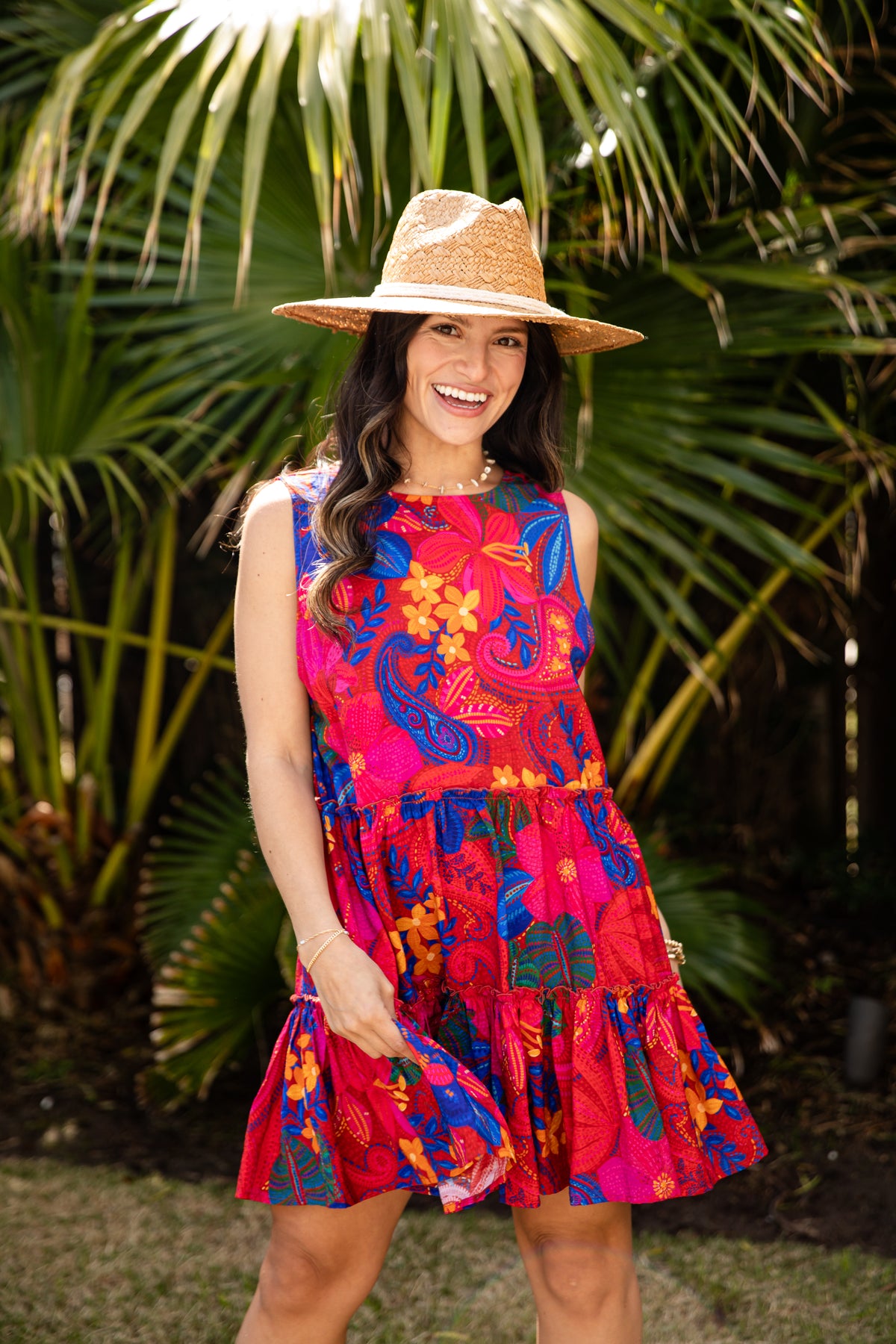 Woman wearing a colorful dress and straw hat standing in front of palm trees.