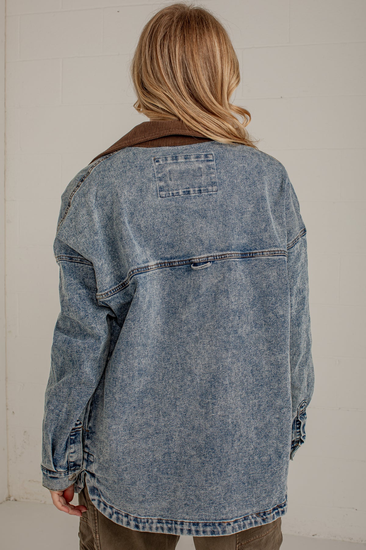 A person viewed from behind wearing an oversized washed blue denim jacket with a brown corduroy collar, paired with olive pants, against a light off-white brick wall.