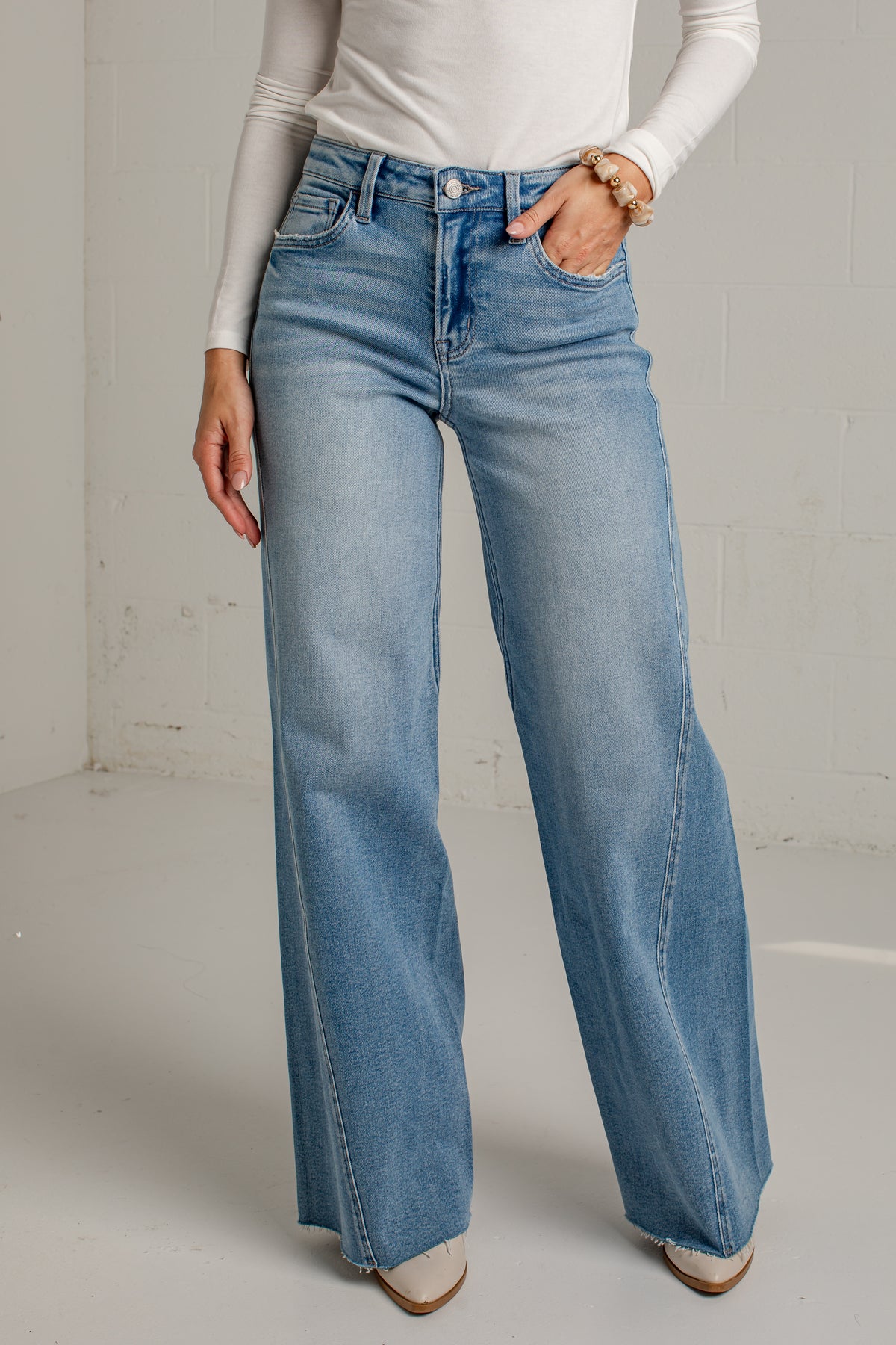 A person wearing light-wash blue wide-leg jeans with a white long-sleeve top, standing in a bright indoor setting with a white wall and floor.
