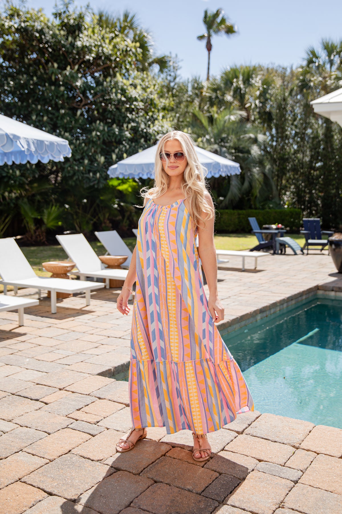 Woman in a colorful dress standing by a pool with lounge chairs and umbrellas in the background.