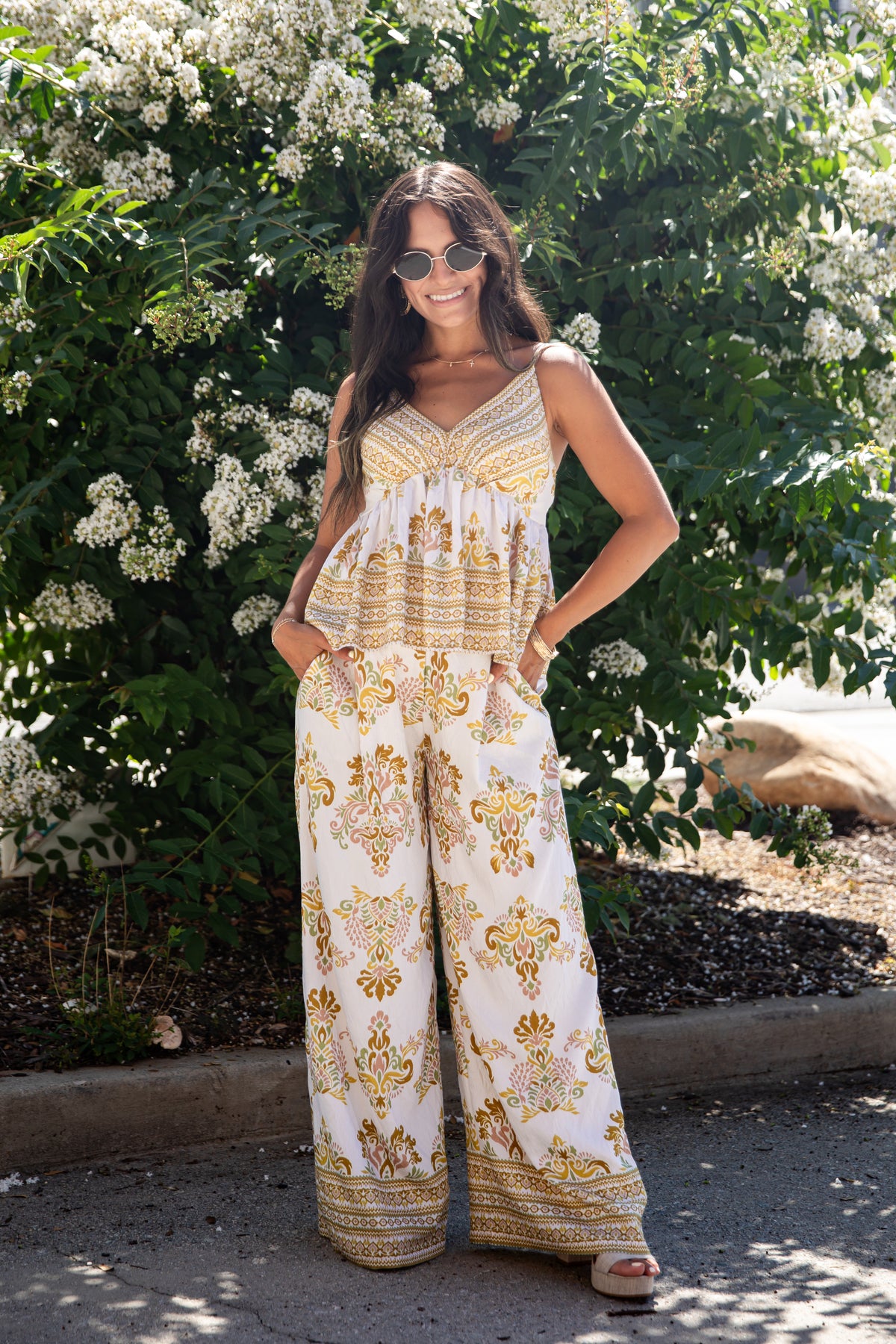 A woman wearing a sleeveless bohemian-style jumpsuit with gold and brown patterns on a white background, standing outdoors in front of a leafy bush with white flowers.