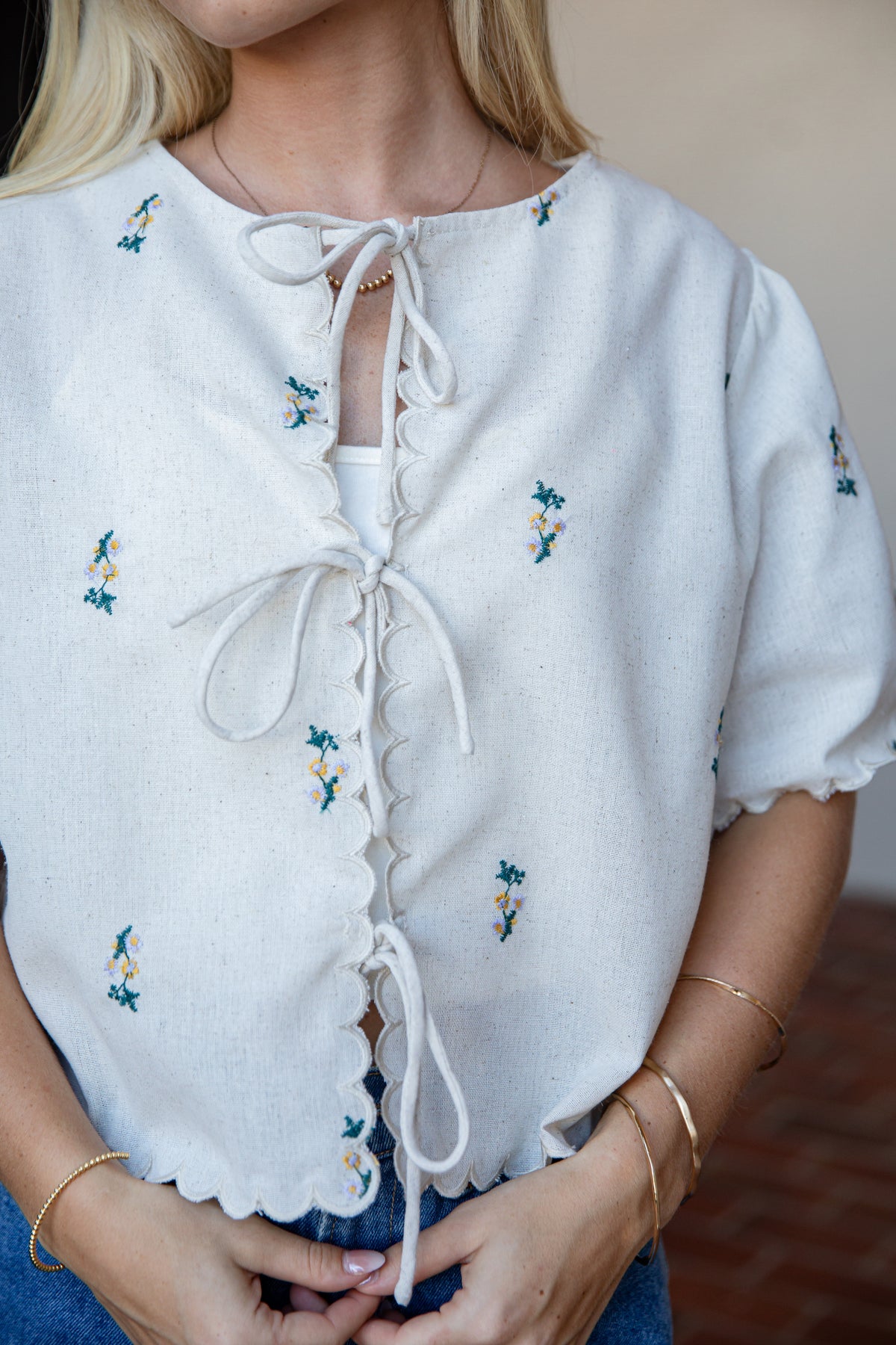 A close-up of a woman wearing a white short-sleeve blouse with small blue and yellow floral embroidery, a scalloped neckline, and front tie closures.