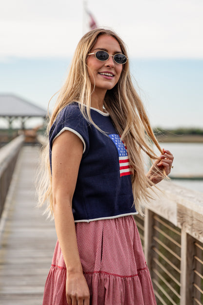 Woman wearing a navy blue t-shirt with an American flag design and a red checkered skirt on a wooden boardwalk.