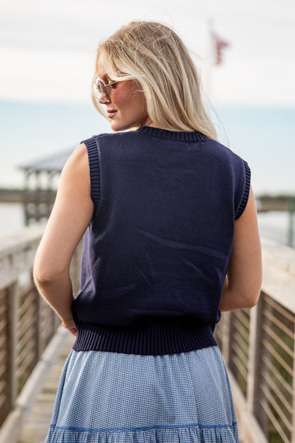 Woman wearing a navy sleeveless sweater and blue skirt on a wooden deck by a body of water.