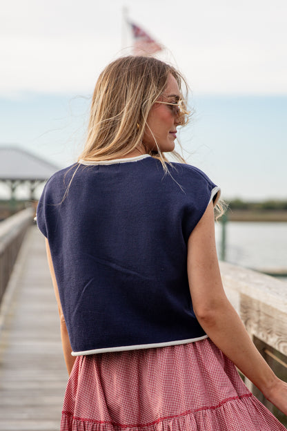 Woman wearing a navy top and red checkered skirt on a wooden dock by water.