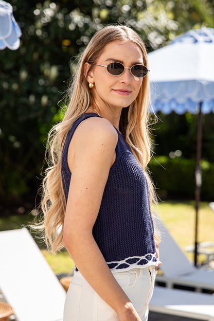 Woman wearing a navy blue sleeveless top and sunglasses outdoors with greenery and an umbrella in the background.