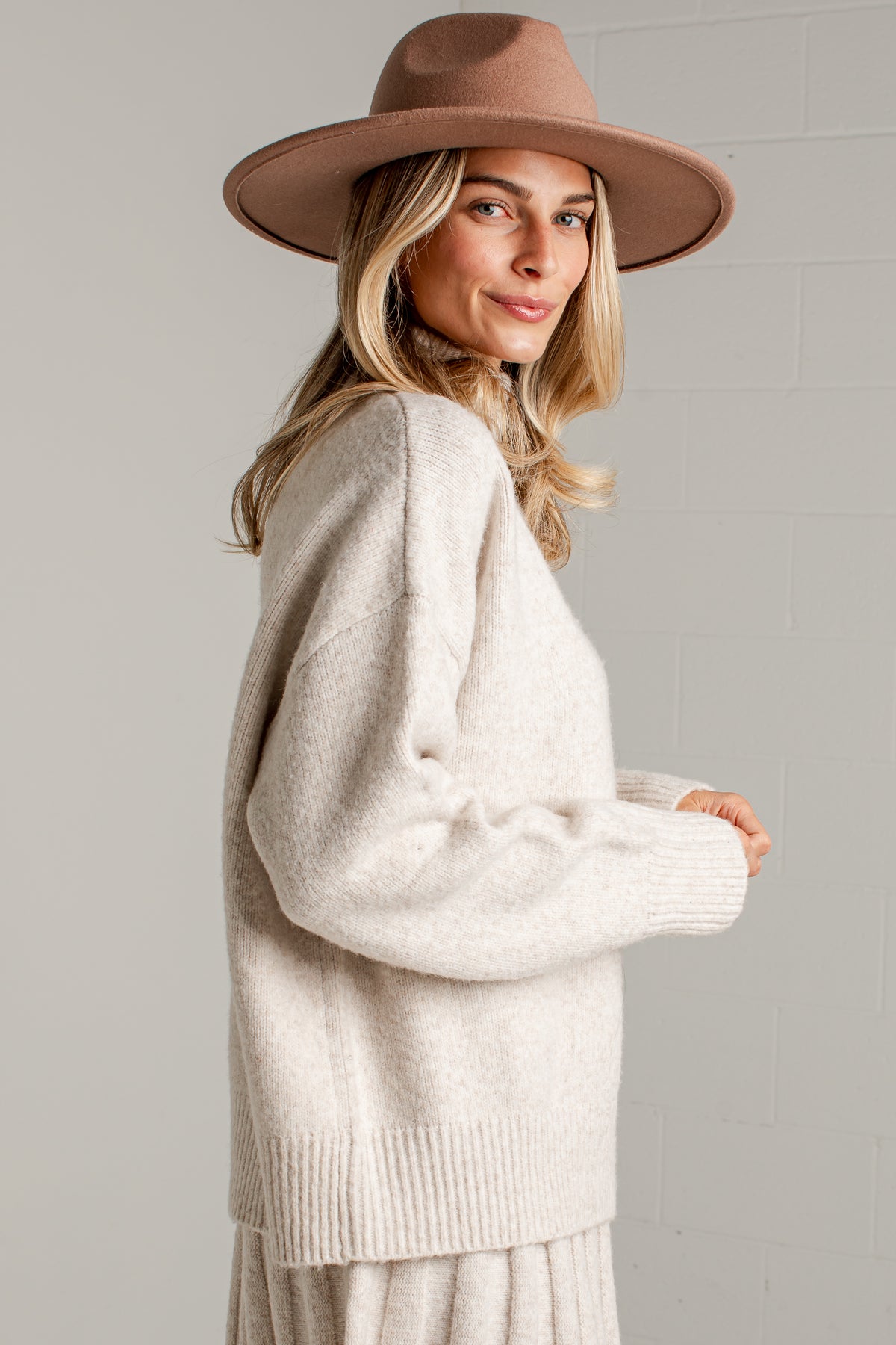 A woman wearing a cream-colored knit sweater and a brown wide-brim hat, photographed against a light gray brick wall.