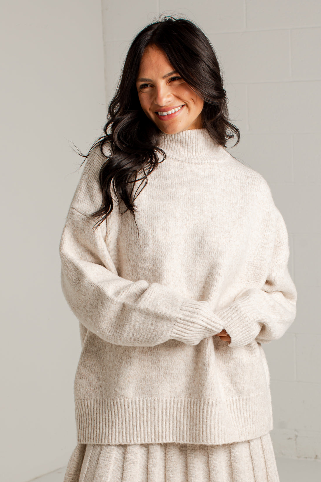 A woman wearing an oversized cream-colored knit mock neck sweater, standing against a light gray tiled wall.