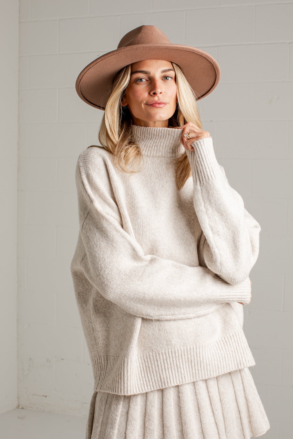 A woman wearing a cream-colored ribbed turtleneck sweater, a matching pleated skirt, and a brown wide-brim fedora hat, standing against a white brick wall.