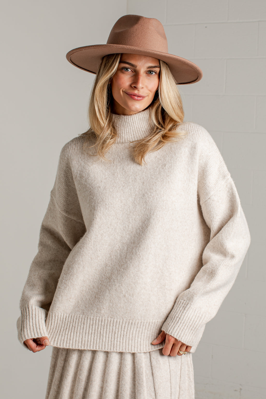 A woman wearing a cream-colored knit mock neck sweater, a matching pleated skirt, and a brown wide-brim fedora hat, standing against a light gray brick wall.