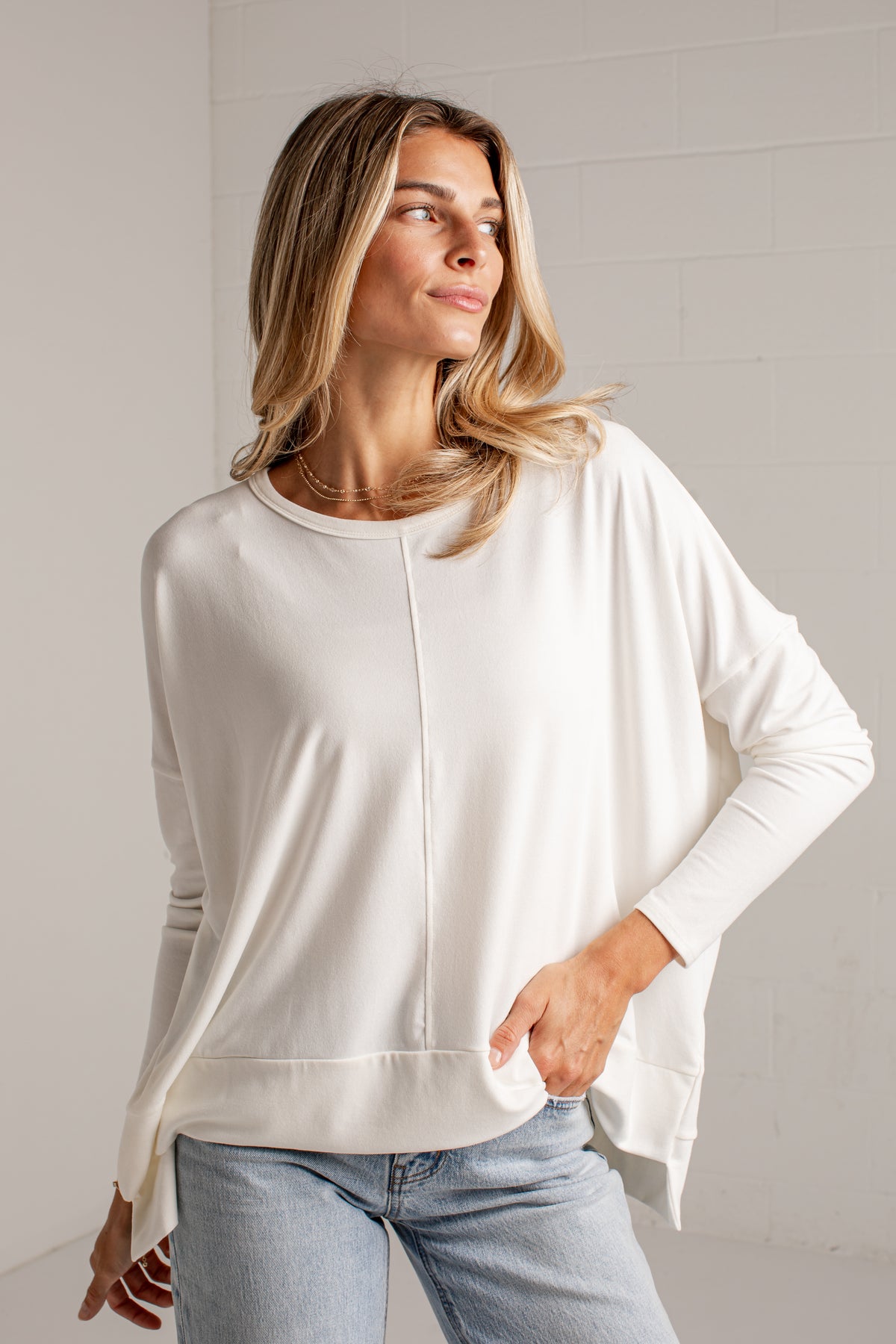 A woman wearing a cream-colored, long-sleeve, relaxed-fit top with a front zipper detail, paired with light-wash blue jeans, standing against a light gray brick wall.