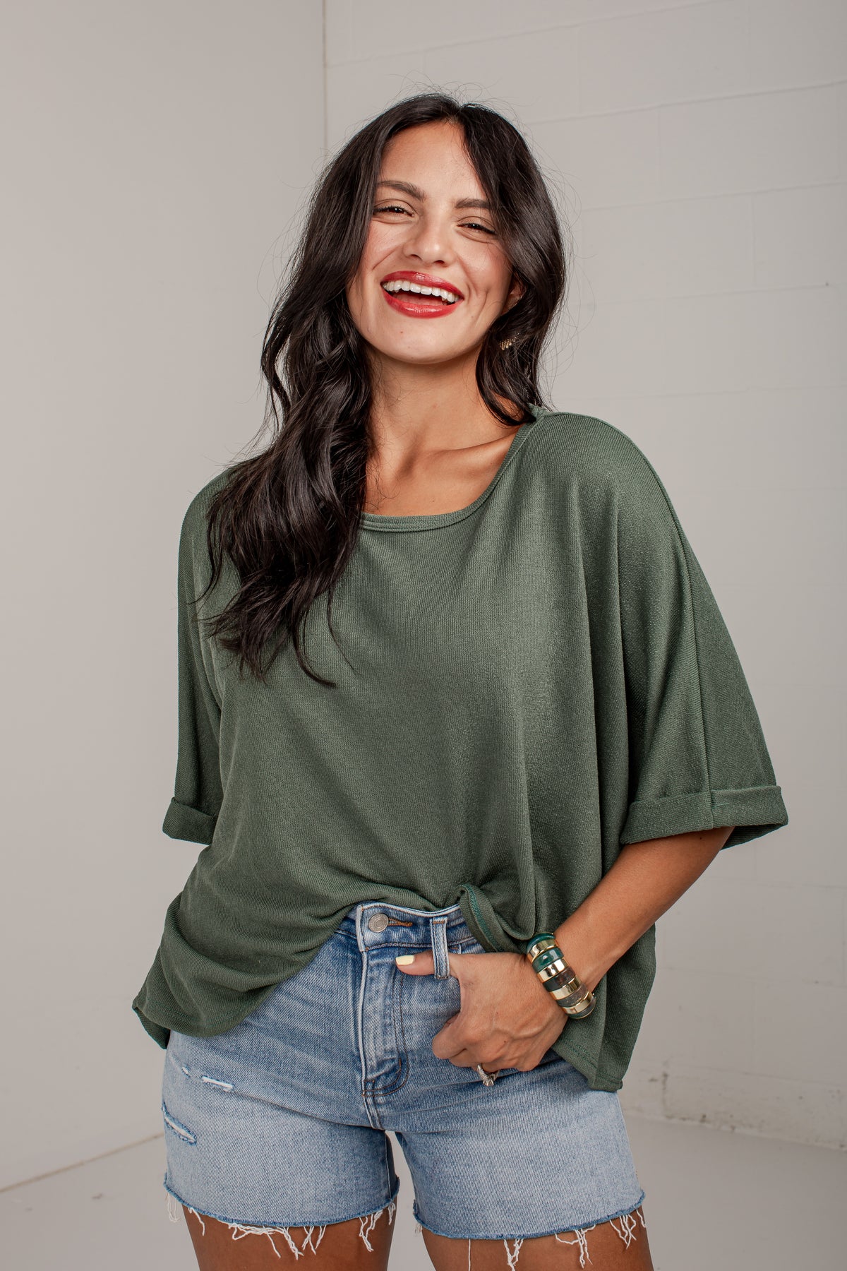 A woman smiling and wearing an olive green short-sleeve tunic top with light blue denim shorts, standing against a light gray wall.