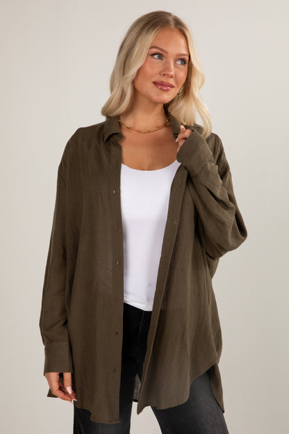 Woman wearing an olive green shirt over a white top and black pants on a plain background