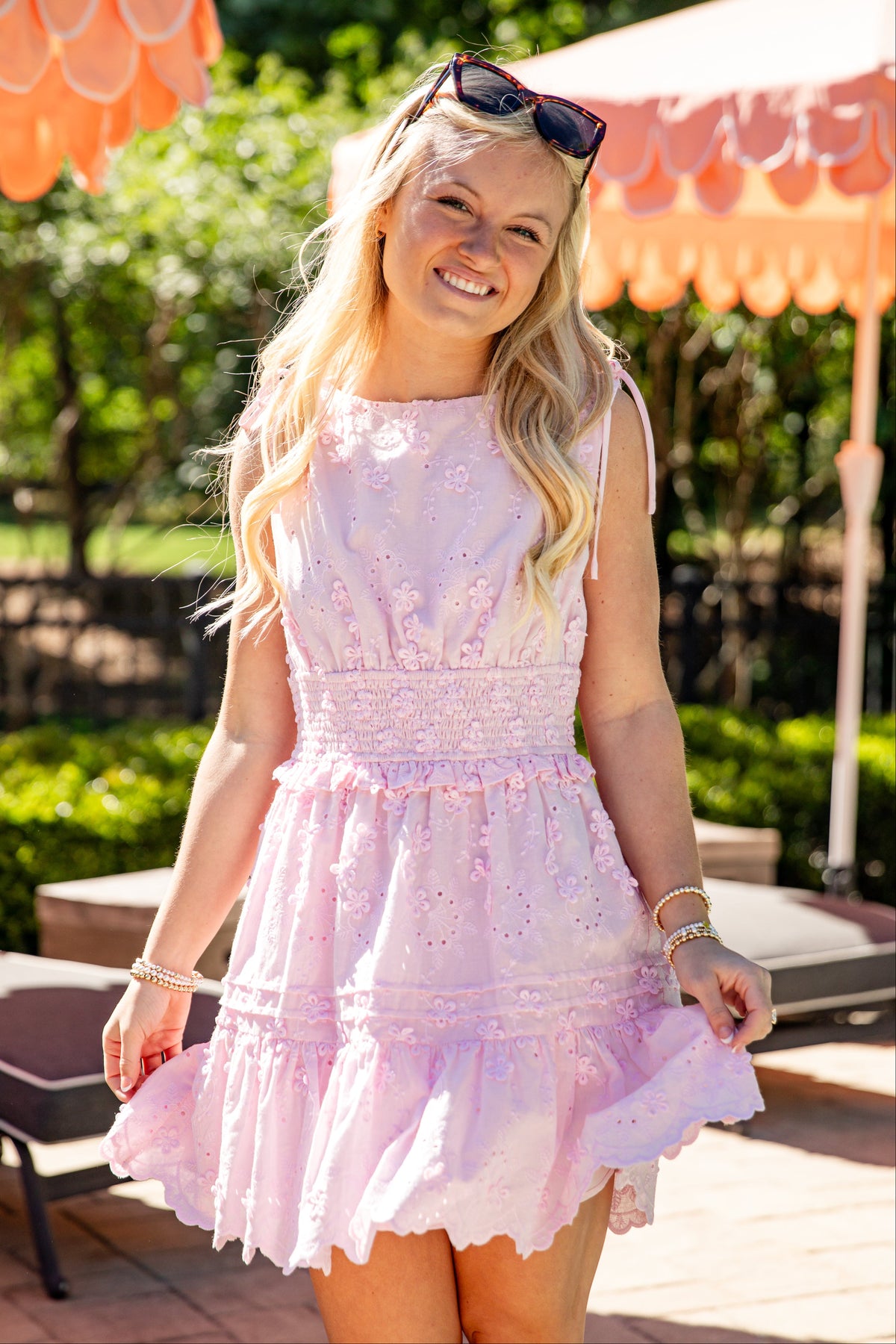 Model wearing a light pink embroidered eyelet mini dress with tie shoulder straps, a smocked waist, and a tiered ruffle skirt.