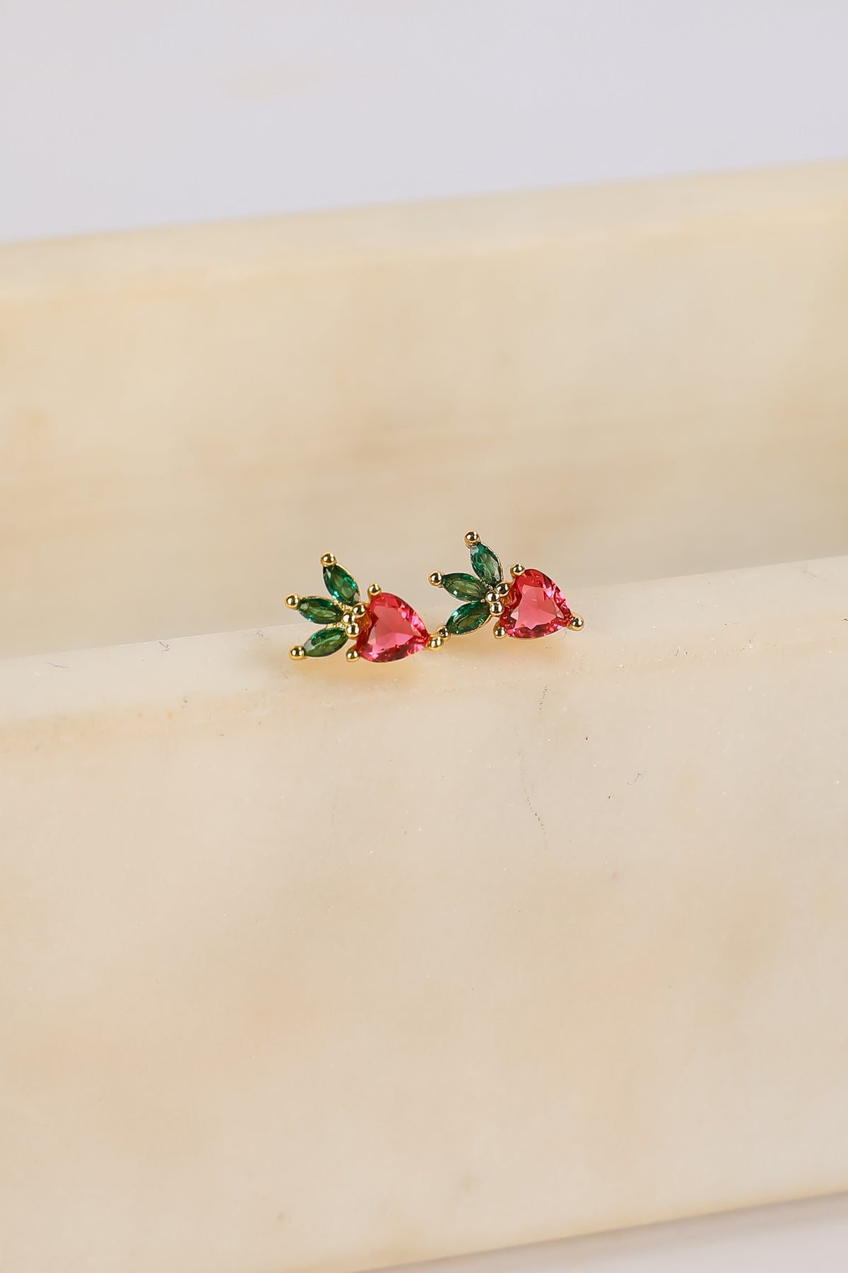 Heart-shaped earrings with green and red stones on a beige surface