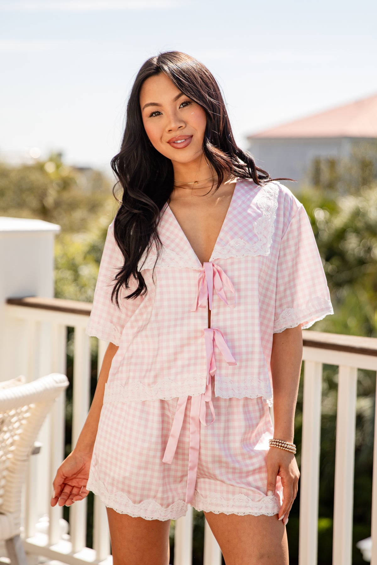 Woman wearing a light pink checkered romper on a balcony with greenery in the background