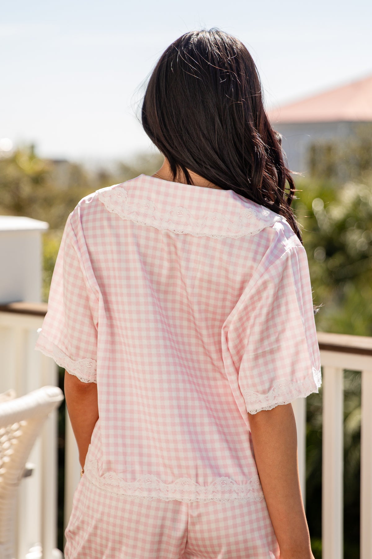 Person wearing a light pink checkered outfit on a balcony with greenery in the background