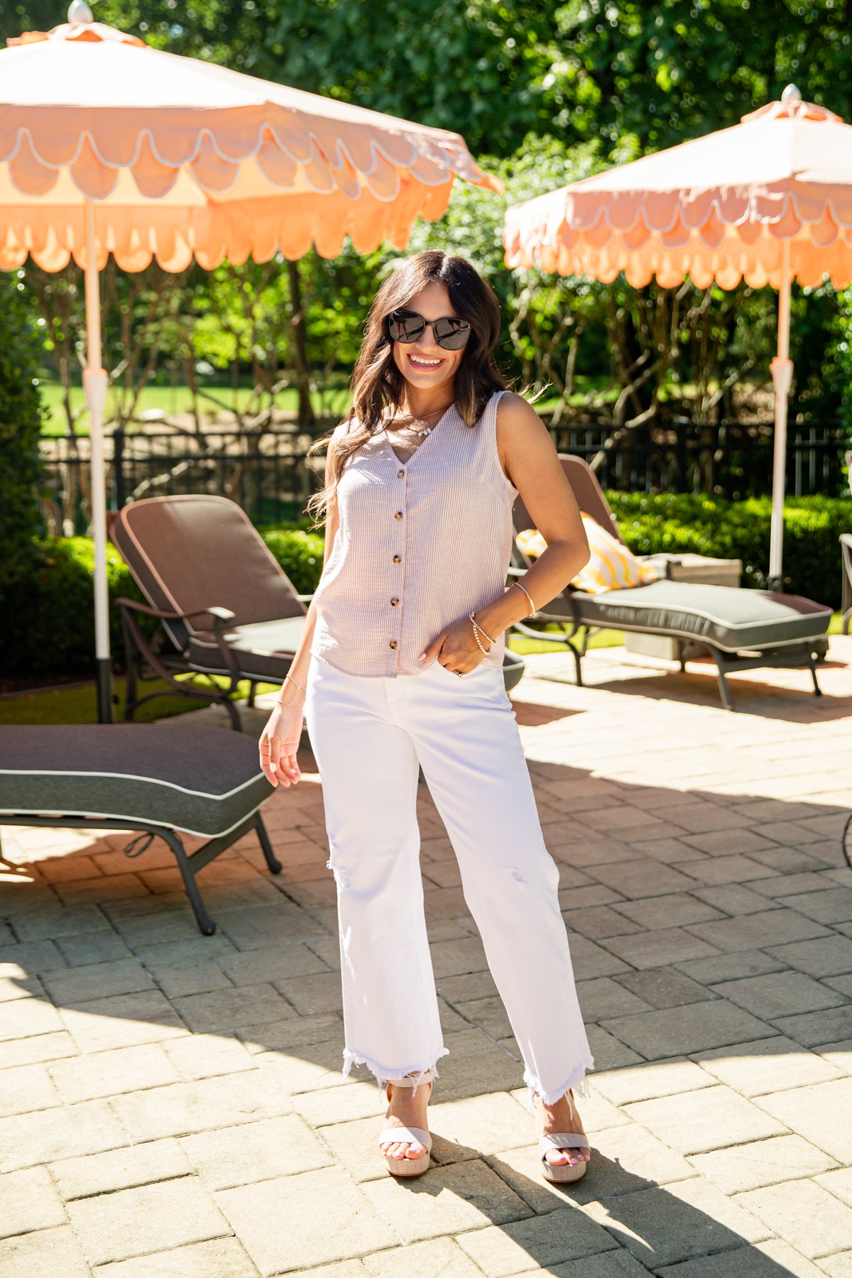 Woman in a light-colored sleeveless top and white pants standing on a patio with orange umbrellas and lounge chairs.
