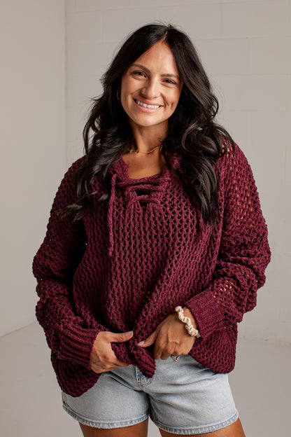 A smiling woman with long dark hair wears an oversized chunky-knit maroon sweater with tie details at the neckline, paired with light-wash denim shorts, standing against a light gray wall.
